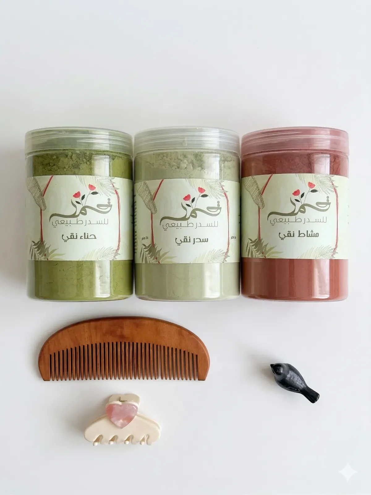 Three Qamar herbal-care jars with botanical labels and a wooden comb.